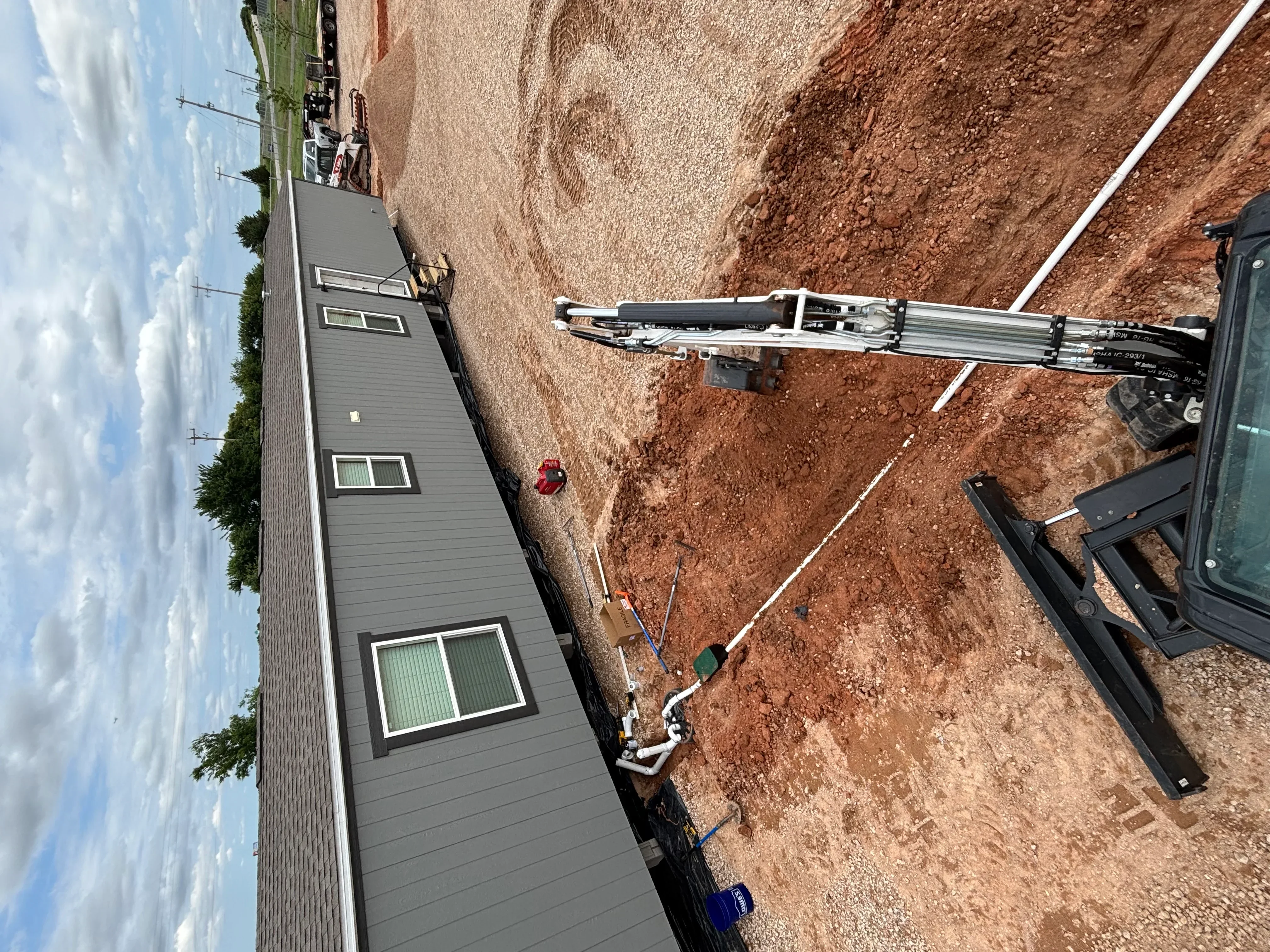 Excavated driveway site with white PVC drainage pipe and compacted clay soil adjacent to gray-sided house
